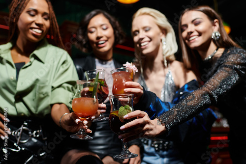 smiling and stylish multiethnic girlfriends clinking cocktails in bar on blurred background