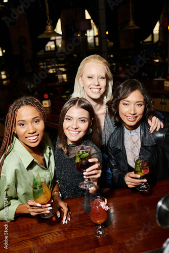 trendy and happy multiethnic female friends holding delicious cocktails and looking at camera in bar