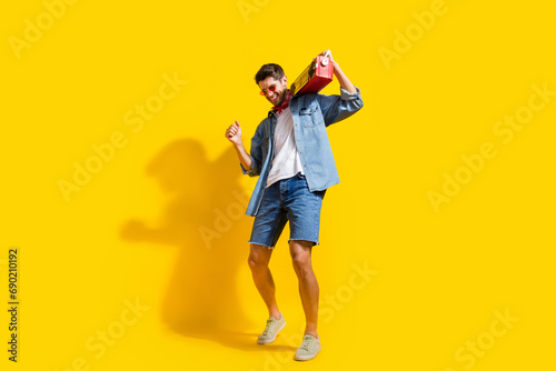 Full body photo of eccentric man dressed denim shirt shorts in glasses dance with boombox on shoulder isolated on yellow color background