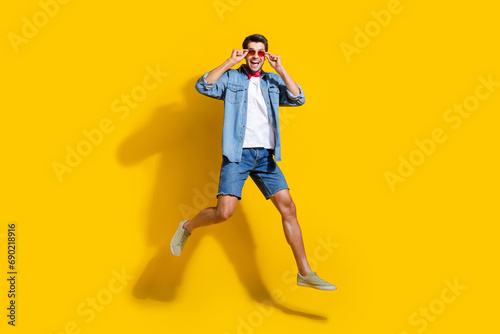 Full body photo of funky optimistic man dressed denim shirt shorts jumping touching glasses isolated on vivid yellow color background.