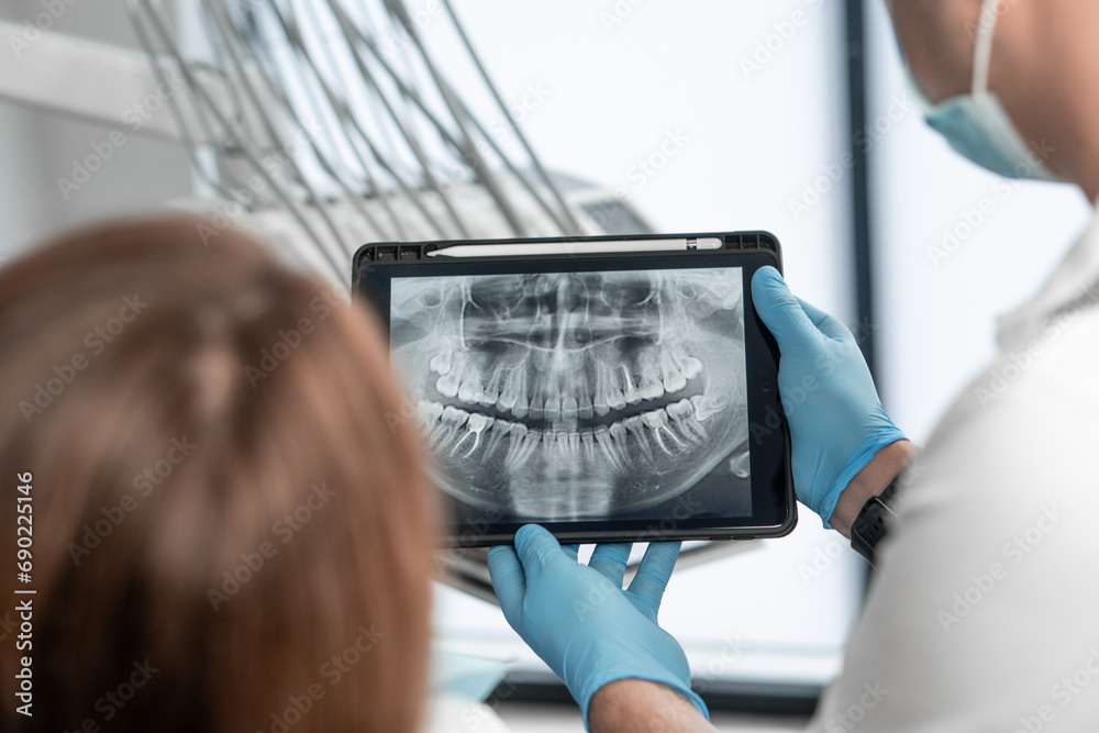 The dentist shows the results of panoramic tomography of the patient's teeth on a tablet. The patient receives an examination, consultation and treatment plan for her oral cavity using an X-ray - obrazy, fototapety, plakaty 