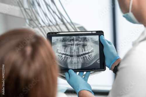 The dentist shows the results of panoramic tomography of the patient's teeth on a tablet. The patient receives an examination, consultation and treatment plan for her oral cavity using an X-ray photo