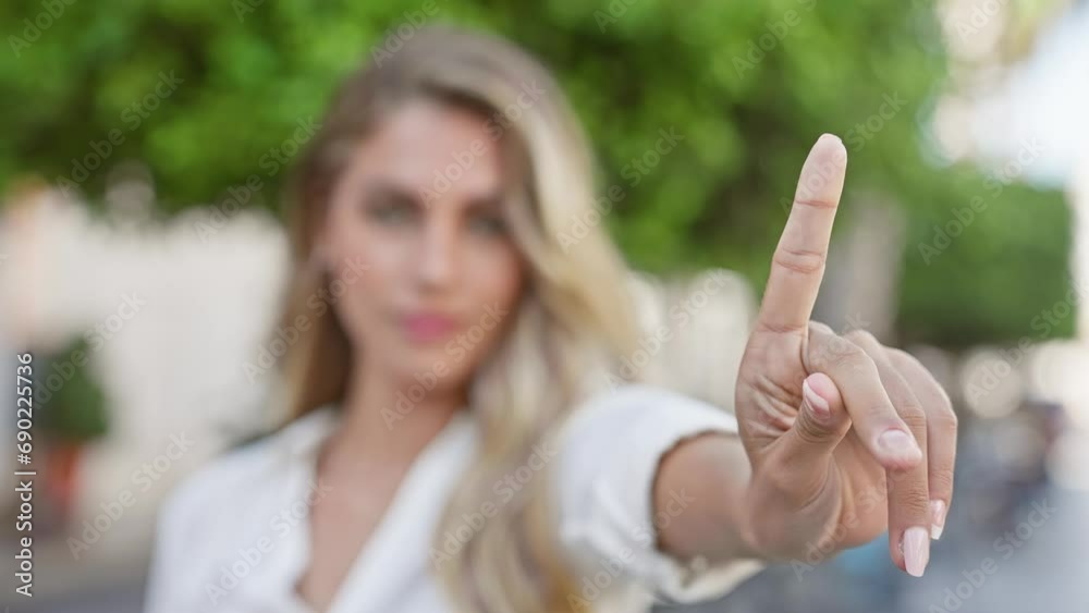 Cool, young blonde woman exudes relaxed, yet serious energy, standing outdoors in sunny, urban street, resolutely saying no with her finger while maintaining eye contact with camera Cool, young blonde woman exudes relaxed, yet serious energy, standing outdoors in sunny, urban street, resolutely saying no with her finger while maintaining eye contact with camera