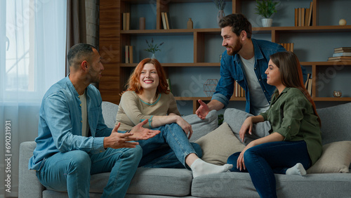 Four ethnic friends fellows companions sit on home couch talking discuss plans chatting friendly talk indoors gathered multiethnic men and women converse joking laughing sharing news real friendship photo