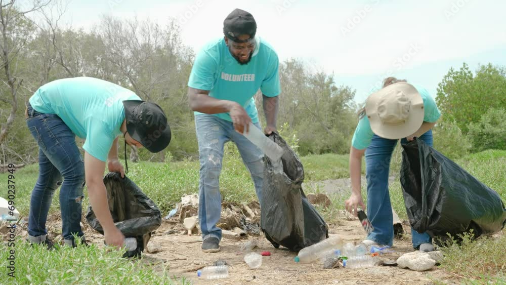 Three volunteers in work overalls collect put garbage in plastic bag at forest. Close-up during action. Cleaning of nature from waste. Environmental assistance. Garbage collection in forest Three volunteers in work overalls collect put garbage in plastic bag at forest. Close-up during action. Cleaning of nature from waste. Environmental assistance. Garbage collection in forest
