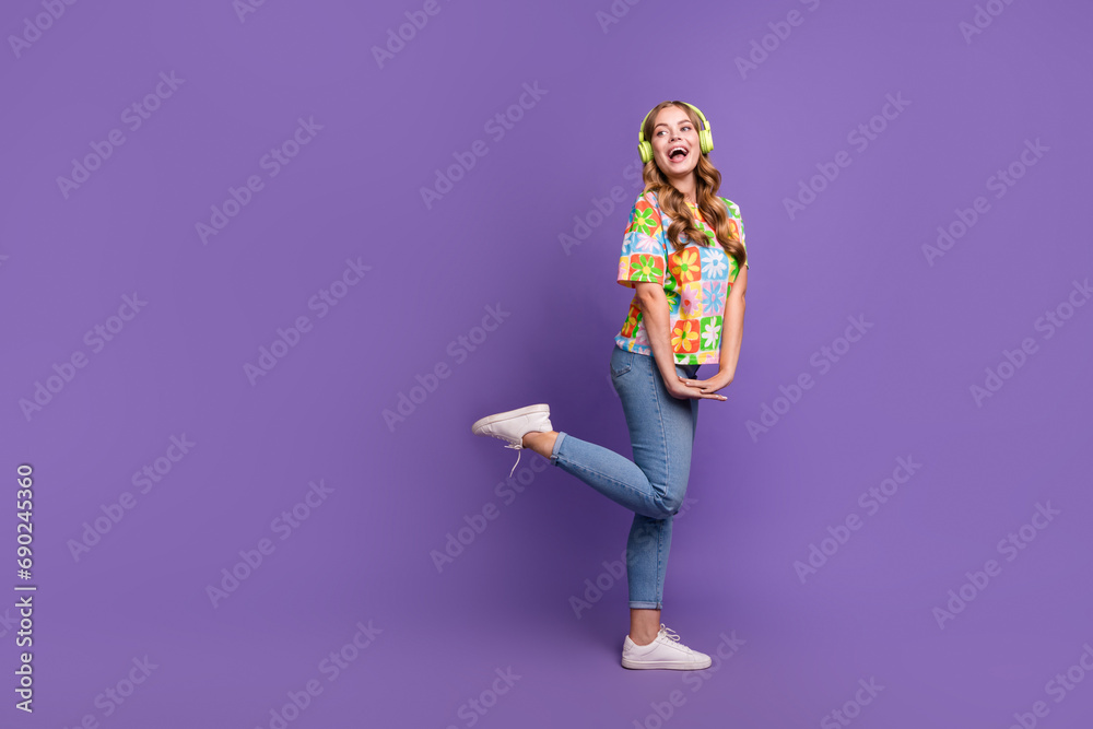 Full size photo of good mood girl dressed print t-shirt jeans in headphones look at promo empty space isolated on violet color background