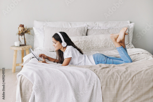 Adorable pre-teen girl in wireless headphones lying on bed with digital tablet device