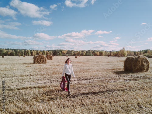 Woman holding colourful shawl walks on countryside field against blue sky. Tourist enjoys exploring and uniting with autumn nature