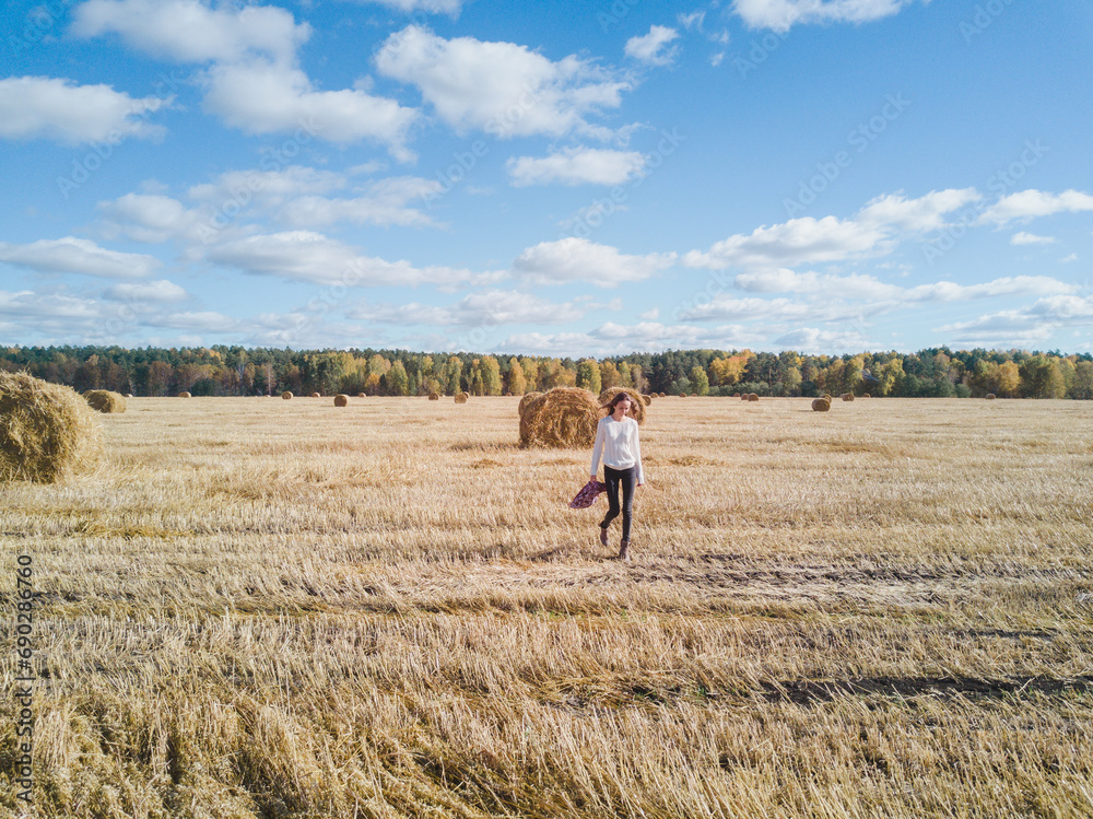 Woman enjoys walking and spending weekend on harvested farm field under blue sky. Tourist explores field with straw bales against forest