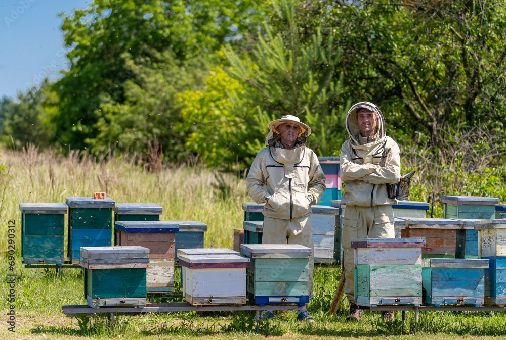 Two men in bee suits standing in front of beehives. Two Beekeepers Inspecting Beehives in Protective Suits Two men in bee suits standing in front of beehives. Two Beekeepers Inspecting Beehives in Protective Suits