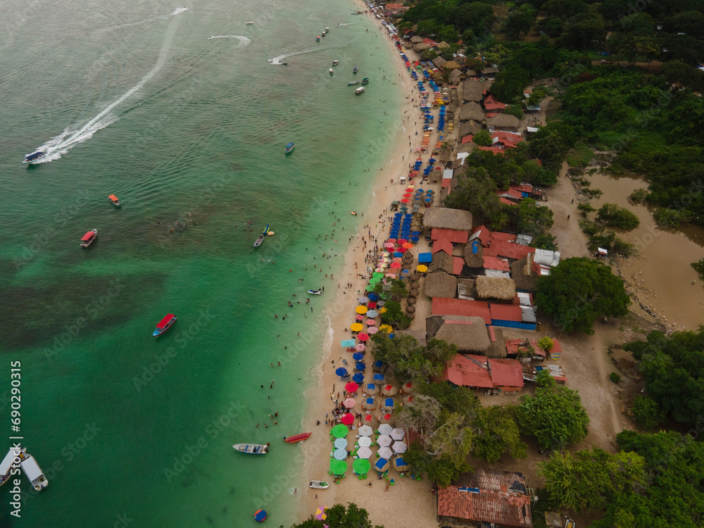 Aerial drone view from above of a small tourist coastal town crowded beach with colorful sun umbrellas, clear ocean water, people swimming, moored boats.