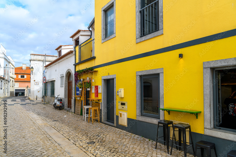 A small, brightly yellow painted sidewalk cafe with barstools in the historic center of the seaside resort town of Cascais, Portugal.