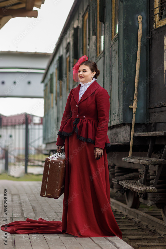 Fototapeta premium A beautiful girl in a burgundy suit of the last century and a hat with a veil stands with a suitcase in her hands near an old steam locomotive.Vintage portrait of the last century, retro journey