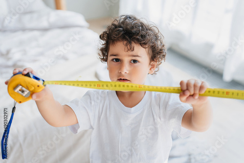Curly confused handsome little boy holding measuring roulette, wants growing faster. Children psychology. Pretty kid puzzled by his small height. Spanish kid with tape measuring at home showing height photo