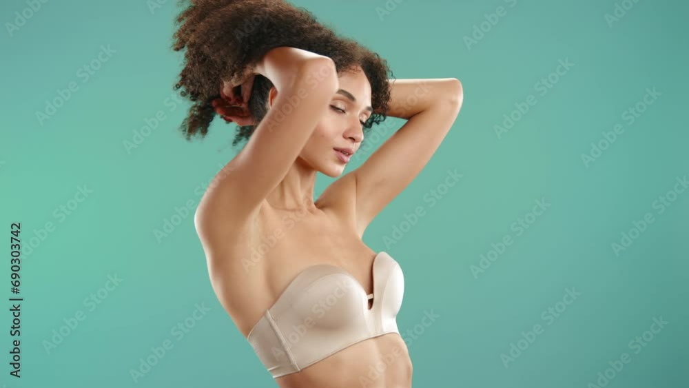 Attractive young model with beautiful body wearing ultimate strapless nude bra closing eyes, raising dark fluffy curls posing on blue green background. High quality 4k footage