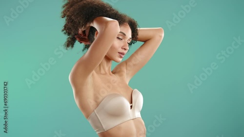 Attractive young model with beautiful body wearing ultimate strapless nude bra closing eyes, raising dark fluffy curls posing on blue green background. High quality 4k footage