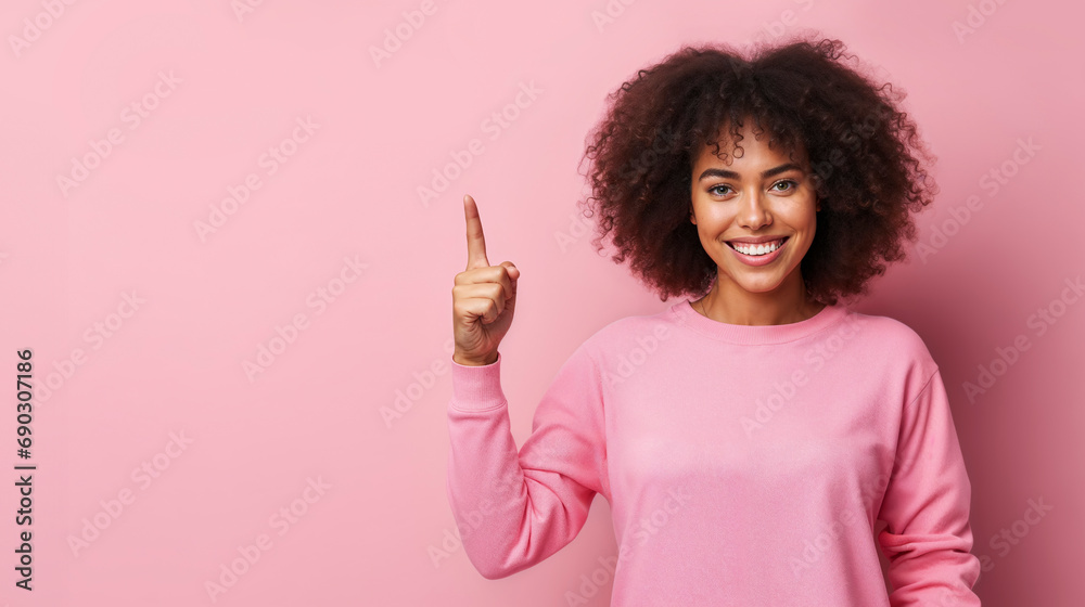 Enthusiastic young woman with curly hair, wearing a pink sweater and pointing upwards, with a bright smile on a pink background. Enthusiastic young woman with curly hair, wearing a pink sweater and pointing upwards, with a bright smile on a pink background.
