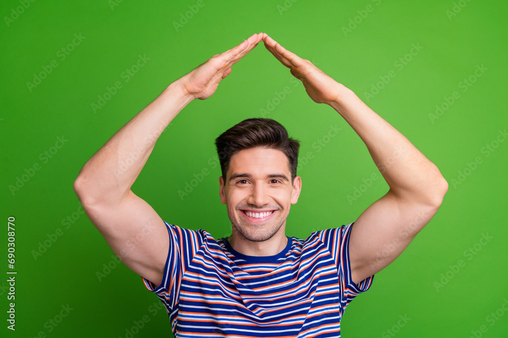 Fototapeta premium Photo of toothy beaming nice guy with stubble dressed striped t-shirt make roof with hands over head isolated on green color background