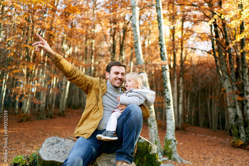 Smiling dad with a little girl on his knees sits on a stump and points his finger at a tree