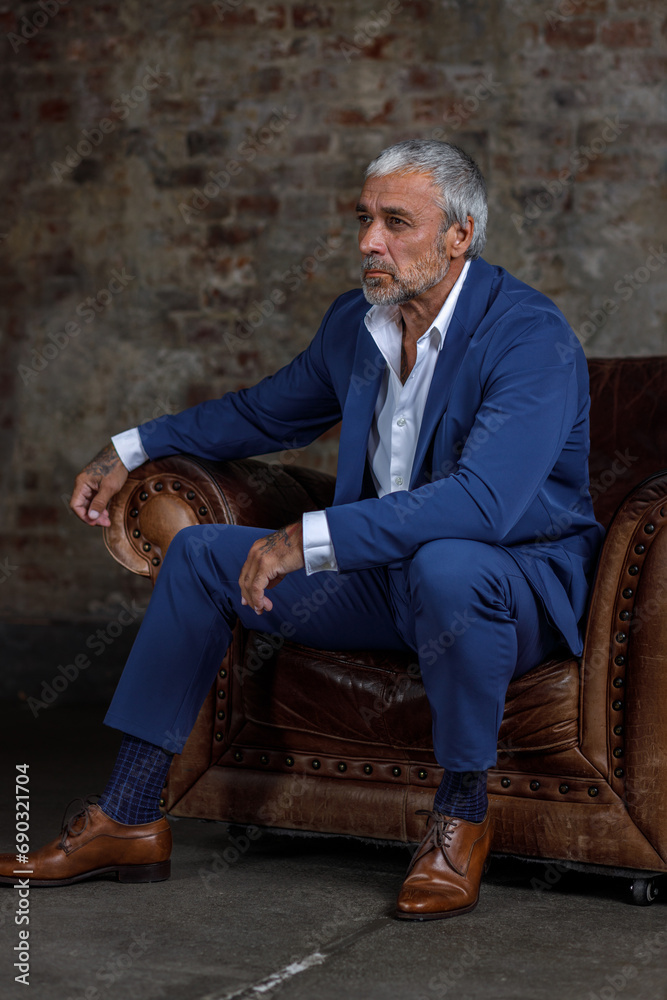 A handsome middle-aged man in a blue formal suit poses sitting on a leather armchair. Portrait of a businessman. A handsome middle-aged man poses on a brick background with a place to copy A handsome middle-aged man in a blue formal suit poses sitting on a leather armchair. Portrait of a businessman. A handsome middle-aged man poses on a brick background with a place to copy