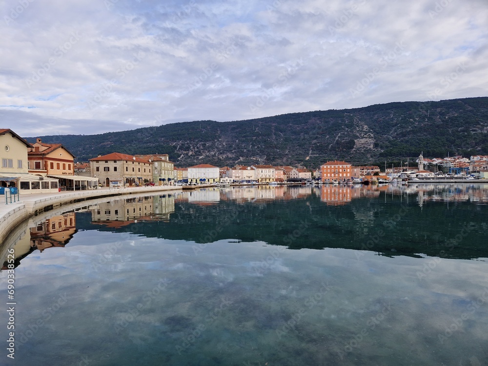 Obraz premium Harbour of Cres on a sunny day in autumn, blue sky, green hills, Croatia island.Sky and houses reflected on the calm sea in Cres gulf