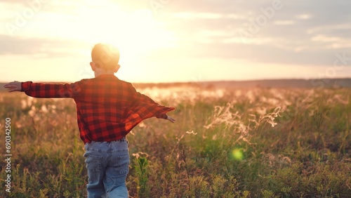 Positive boy runs along field with growing plants at sunset in autumn evening. Boy runs imitating flight with arms out to sides in rural nature with agriculture. Boy feels freedom running