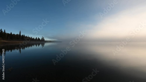 Time lapse of a calm northern lake in the morning as fog moves across the water.  The water reflects both the fog and the blue sky. A point of land projects from the left side and is covered with ever photo