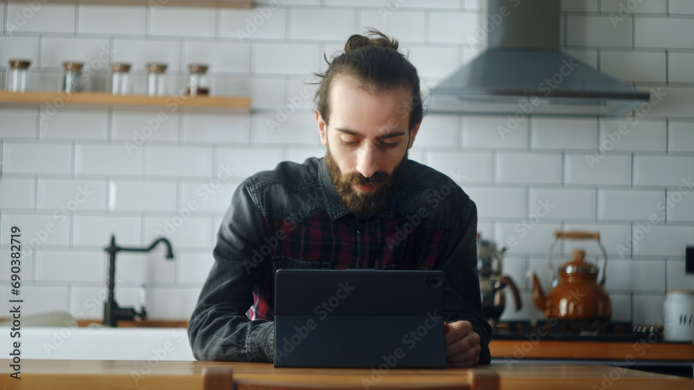 Bearded young man sitting in kitchen, using app with tablet device, surfing on internet Bearded young man sitting in kitchen, using app with tablet device, surfing on internet