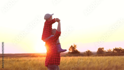 Farmer dad carries infant boy on shoulders. Farmer dad wanders through wheat field with child sitting atop shoulders. In expanse of wheat farmer family delights in stroll together at sunset on horizon