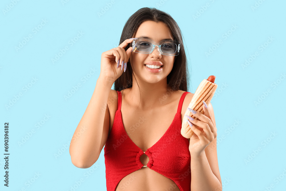 Pretty young woman in swimsuit and with tasty french hot dog on blue background Pretty young woman in swimsuit and with tasty french hot dog on blue background