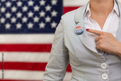 Young voter woman  waist up pointing with finger to usa badge at vote center. Woman stands U.S. flag in background at electoral college presidential election day. Motivation of a political campaign. 