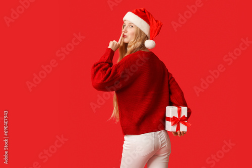 Beautiful young woman in Santa hat and warm winter clothes with gift box showing silent gesture on red background