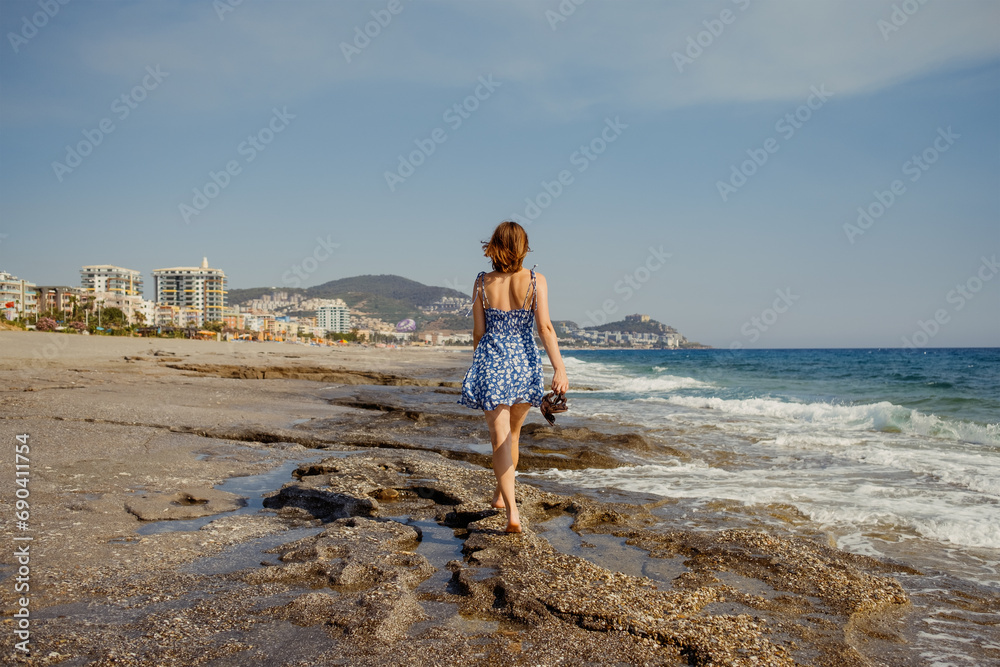 A lone wanderer traverses the rocky beach, with the sea breeze guiding her steps and the cityscape in the distance