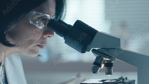 Senior female chemist in protective eyewear and gloves pouring blood sample on stage of microscope and observing it at work in laboratory photo