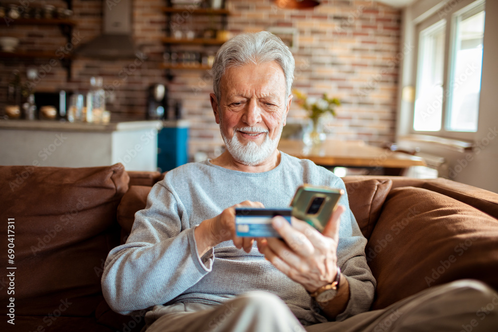 Senior man sitting on sofa with smartphone using credit card at home Senior man sitting on sofa with smartphone using credit card at home