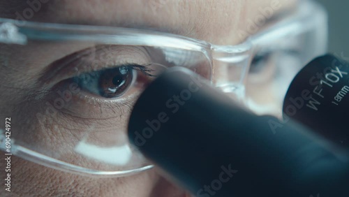 Extreme close-up shot of eyes of mature female scientist in protective glasses looking through microscope eyepiece at work in laboratory