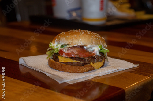 hamburguesa con queso, tomate y lechuga, sobre una servilleta blanca en un restaurante de comida rápida con bebidas de fondo