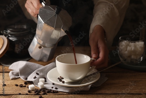 Tableau sur toile Woman pouring aromatic coffee from moka pot into cup at wooden table, closeup