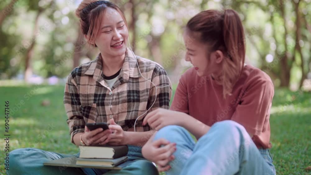 Asian female college student is using her smartphone to listen to music through headphones and sharing the experience with her friend, aiming to unwind and relax in a garden with lush green trees. Asian female college student is using her smartphone to listen to music through headphones and sharing the experience with her friend, aiming to unwind and relax in a garden with lush green trees.
