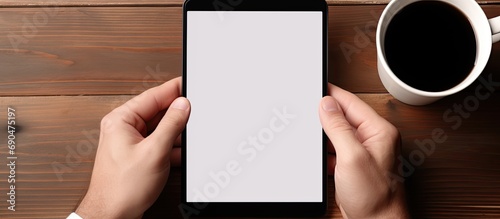 Man's hands holding a black blank screen smartphone, sitting in front of a computer tablet with a white blank screen and stylus pen, on a wooden working desk.