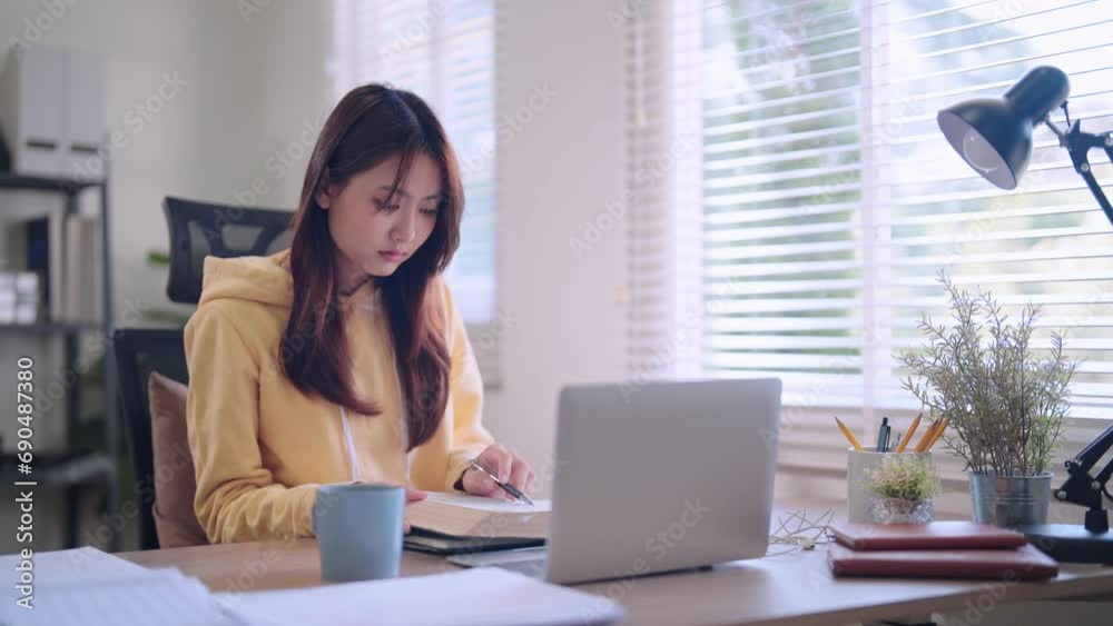 Asian female student engages in focused online learning using her laptop and headphones. She diligently records study materials in a notebook at her home workspace. Asian female student engages in focused online learning using her laptop and headphones. She diligently records study materials in a notebook at her home workspace.