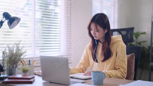 Asian female student engaged in online learning using a laptop and headphones, diligently recording study materials in a somewhat bored manner at her home desk photo