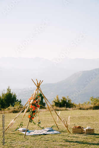 Tepee-style wedding arch stands over a blanket and pillows next to food crates on the lawn