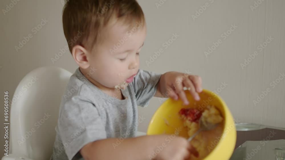 A cute toddler boy eating a healthy breakfast of cereal in the kitchen in the morning. A baby learning to eat with a spoon. Complementary feeding are new stage of feeding of child. A cute toddler boy eating a healthy breakfast of cereal in the kitchen in the morning. A baby learning to eat with a spoon. Complementary feeding are new stage of feeding of child.