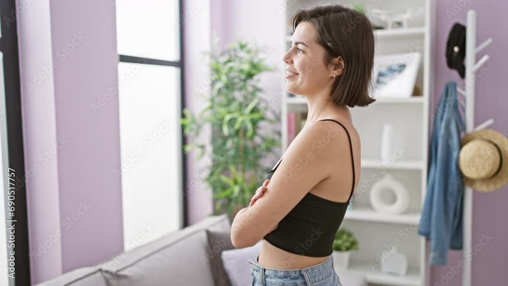 Joyful hispanic woman smiling while standing in her living room, performing a relaxed arms-crossed gesture, gazing through the window with confidence Joyful hispanic woman smiling while standing in her living room, performing a relaxed arms-crossed gesture, gazing through the window with confidence