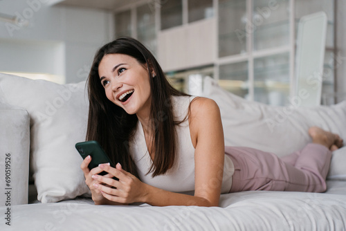Brunette cheerful girl laying on couch at home using phone laughing watching funny videos on social media feed. Weekend, female relaxing indoors.
