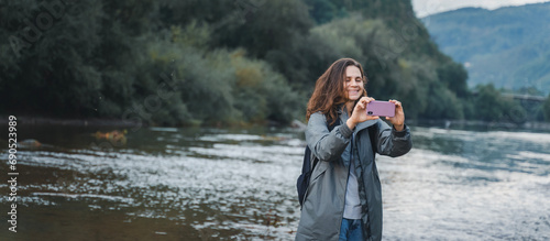 A young woman traveler in a raincoat with a smartphone in her hands on the bank of a mountain rive