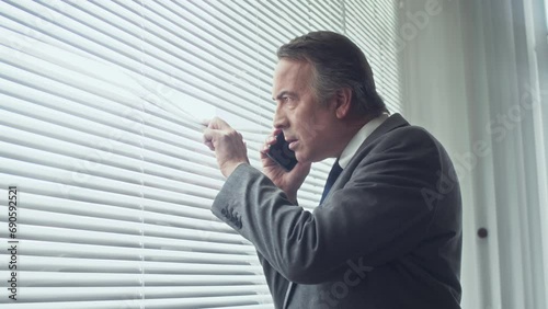 Side view of senior businessman suspecting someone while talking on smartphone looking out window photo