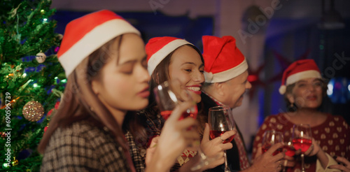 Indian Asian young adult older lady staff male group teen girl friend santa hat cap indoor office drink wine juice happy old aged man people hold glasses make fun joy new year 2024 enjoy night party