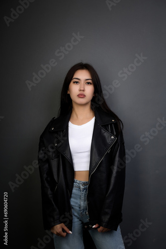 Serious calm Asian girl looking directly at the camera on a gray background. Asian model in jeans, white T-shirt and leather jacket posing for camera for portfolio